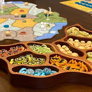 May include: A board game with a colorful map and wooden game pieces. A wooden organizer holds various game tokens in different colors and shapes. The game board features a landscape with land and water, and small wooden figures.