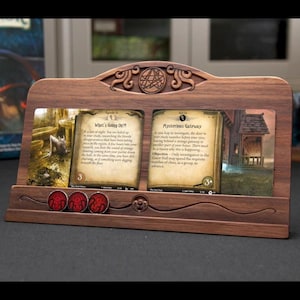 May include: A dark wood card display stand featuring an arched top with a circular engraved symbol. The stand holds two game cards titled What's Going On?! and Mysterious Gateway, with three circular red tokens placed in a horizontal groove below.
