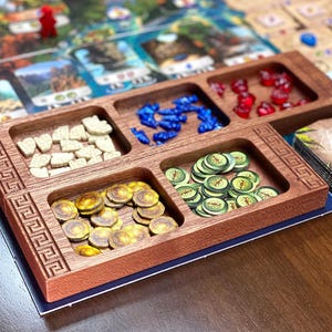 May include: A wooden game organiser with compartments holding game pieces. The pieces are various colours and shapes, including gold coins, blue gems, and green tokens. The organiser sits on a game board.