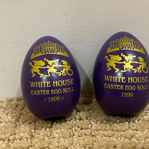 Puede incluir: Dos huevos de Pascua morados con letras doradas y una imagen dorada de la Casa Blanca. Los huevos dicen "White House Easter Egg Roll 1999".