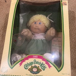 May include: A Cabbage Patch Kid doll in a green dress and white shoes. The doll has blonde hair and blue eyes. The doll is in a box with the words "Cabbage Patch Kids" and "I'm unique. I'm adoptable!" on it. The box also has the year 1985 printed on it.