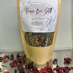 May include: A brown paper bag with a white label that reads "Peace Be Still Tisane Blend" and lists the ingredients as chamomile, holy basil, hibiscus, lemon balm, and peppermint. The bag is filled with loose leaf tea.