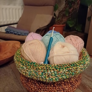 Könnte beinhalten: Ein handgefertigter Häkelkorb gefüllt mit verschiedenfarbigen Wollknäueln und einem Häkelhaken. Der Korb ist zweifarbig, mit einem braunen Boden und einem grünen, gelben und braunen Rand. Die Garnfarben sind rosa, hellblau und creme.