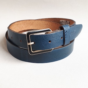 以下が含まれることがあります： 銀と黒のバックルが付いた濃い青色の革ベルト。ベルトは巻かれており、バックルが見えています。
