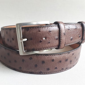 May include: A brown leather belt with a silver buckle. The belt has a textured surface with small, dark spots. The buckle is square and made of shiny metal. The belt is rolled up on a white surface.