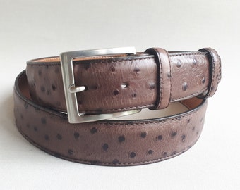 May include: A brown leather belt with a silver buckle. The belt has a textured surface with small, dark spots. The buckle is square and made of shiny metal. The belt is rolled up on a white surface.