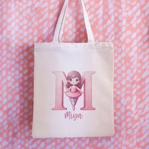 Peut inclure: Un sac fourre-tout en toile blanche avec une lettre M rose et une illustration de ballerine. Le nom "Miya" est imprimé sous la lettre.