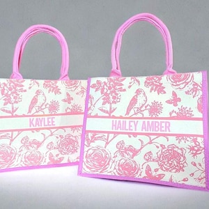 May include: Two pink tote bags with floral patterns and pink handles. The bags have white personalized names on them: "KAYLEE" and "HAILEY AMBER".