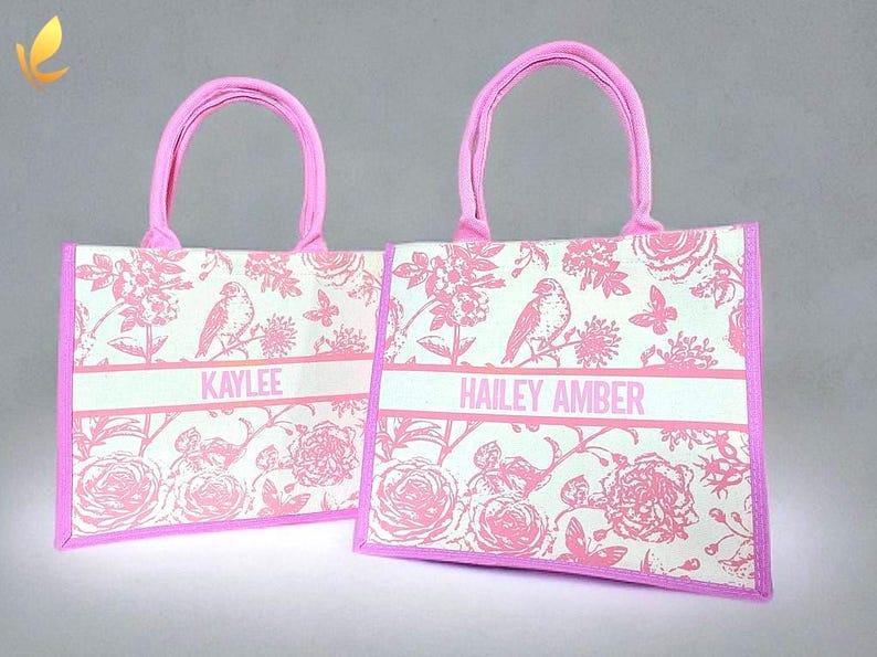 May include: Two pink tote bags with floral patterns and pink handles. The bags have white personalized names on them: "KAYLEE" and "HAILEY AMBER".