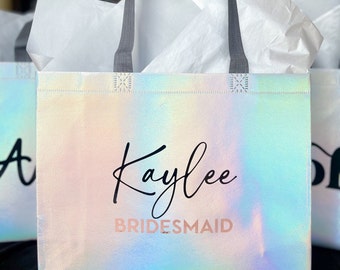 May include: A white iridescent reusable tote bag with gray handles. The bag has the name "Kaylee" written in black cursive script and the word "BRIDESMAID" written in pink below it.