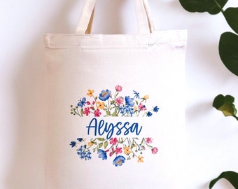 May include: A white canvas tote bag with a floral design and the name "Alyssa" printed in blue. The flowers are blue, pink, yellow, and green.