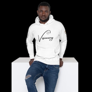 May include: White hooded sweatshirt with a black cursive logo that says "Visionary" and a small circular design on the left side of the chest. The sweatshirt has a pouch pocket and long sleeves.