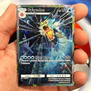 May include: A holographic Gyarados Pokémon card. The card features a blue and white illustration of Gyarados with the text "Gyarados" and "Hyper Beam". The card also includes the HP of 150 and the text "Discard a random Energy from your opponent's Active Pokémon."