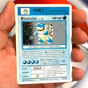 May include: A Blastoise Pokemon card from the 1996 2025 Custom Card set. The card is blue and white with a picture of Blastoise on the front. The card has a HP of 100 and a power called Rain Dance. The card also has a move called Hydro Pump that does 60 damage plus 10 more damage for each Energy attached.