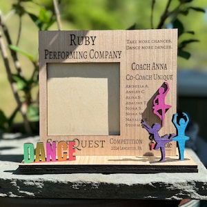 May include: A wooden frame with a photo of dancers in pink, purple, and blue. The frame has the words "Ruby Performing Company" and "Dance Quest Competition 2024 Lancaster, PA" on it.