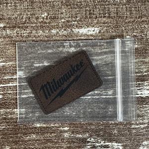 May include: A brown leather patch with the word "Milwaukee" in black lettering and a lightning bolt logo.