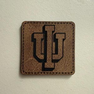 May include: A square, brown leather patch with a stitched border. The patch features the interlocking letters "IU" in black, representing Indiana University. The leather has a textured appearance.