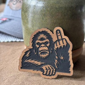 May include: A brown and black patch featuring a Bigfoot-like creature giving the middle finger. The patch has a textured appearance and is likely made of leather or a similar material. The design is a bold, humorous statement.