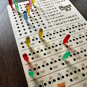 Op de afbeelding: Een houten spelbord met een raster van cirkels en nummers. Het bord is gelabeld "Phase 10" en heeft een lijst met scorevereisten. Gekleurde pinnen worden in de cirkels op het bord geplaatst.