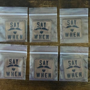 May include: Six small, clear plastic bags containing brown patches with a black mustache, heart, and the words "SAY WHEN." The patches are square with rounded corners and are sealed inside the bags.