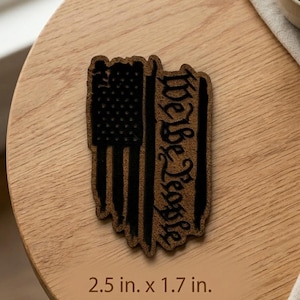 May include: A brown and black patch with the text "We the People" and a distressed American flag design. The patch measures 2.5 inches by 1.7 inches.