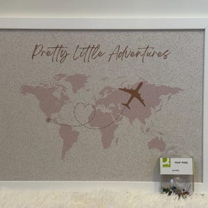 May include: A white framed corkboard with a world map printed on it. The map is pink and has a heart-shaped route drawn on it. The text "Pretty Little Adventures" is printed at the top of the board. A small package of map pins is in the bottom right corner of the image.
