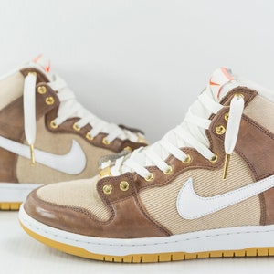May include: A pair of high-top trainers with a brown leather and canvas upper, white swoosh logo, and white laces. The trainers have a white midsole and a gum rubber outsole.