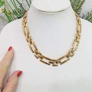 May include: A gold-toned necklace with a geometric design. The necklace is displayed on a white mannequin bust. The necklace features rectangular links connected in a repeating pattern. The jewelry is a statement piece.