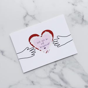 May include: A white card with a red heart and black hand outlines. The heart has a pink paper inside with the text "Will you be my bridesmaid?" written in purple ink.