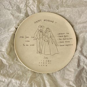 Peut inclure: Une assiette ronde en c&eacute;ramique blanc cass&eacute; avec un dessin au trait noir repr&eacute;sentant un couple de mari&eacute;s. Le texte inclut 'HAPPY WEDDING !!', '10th June 2025', '12 a.m.', 'You are invited.', un menu et un calendrier de juin avec le 4 entour&eacute;.