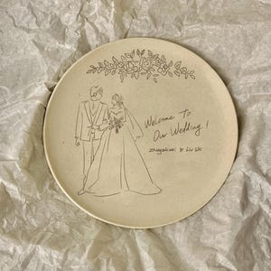 Peut inclure: Une assiette ronde en c&eacute;ramique cr&egrave;me mouchet&eacute;e orn&eacute;e d'une illustration au trait noir repr&eacute;sentant un couple en tenue de mariage. Au-dessus se trouve une guirlande florale, et &agrave; droite le texte "Welcome To Our Wedding!" et "Zhangzhiwei & Liu Shi".