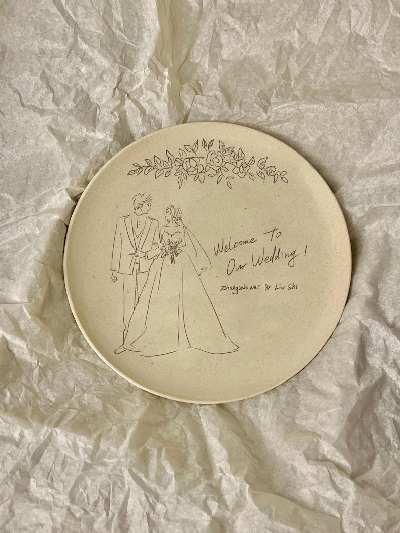 Peut inclure: Une assiette ronde en c&eacute;ramique cr&egrave;me mouchet&eacute;e orn&eacute;e d'une illustration au trait noir repr&eacute;sentant un couple en tenue de mariage. Au-dessus se trouve une guirlande florale, et &agrave; droite le texte "Welcome To Our Wedding!" et "Zhangzhiwei & Liu Shi".