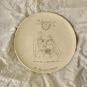 Peut inclure: Une assiette ronde en c&eacute;ramique blanc cass&eacute; mouchet&eacute;e avec une illustration au trait noir repr&eacute;sentant un couple en tenue de mariage. Au-dessus, une couronne florale avec la date 2025 12.25. Texte : MR. AND MRS. LI XUE & ZHOU CHEN et WE ARE GETTING MARRIED!