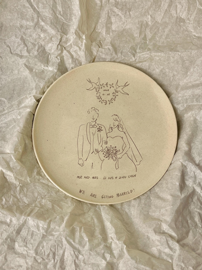 Peut inclure: Une assiette ronde en c&eacute;ramique blanc cass&eacute; mouchet&eacute;e avec une illustration au trait noir repr&eacute;sentant un couple en tenue de mariage. Au-dessus, une couronne florale avec la date 2025 12.25. Texte : MR. AND MRS. LI XUE & ZHOU CHEN et WE ARE GETTING MARRIED!