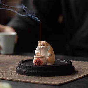 Könnte beinhalten: Ein Keramik-Räucherstäbchenhalter in Form einer Katze, die einen kleinen Topf mit chinesischen Schriftzeichen hält. Die Katze ist orange und weiß und der Topf ist braun mit roten chinesischen Schriftzeichen. Das Räucherstäbchen brennt und Rauch steigt aus der Spitze auf.