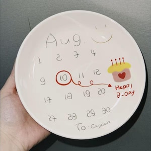May include: A white ceramic plate featuring a hand-drawn calendar design. The month of August is written at the top, with the 10th circled in red. A cupcake with candles and the words "Happy B-Day" are also drawn on the plate.