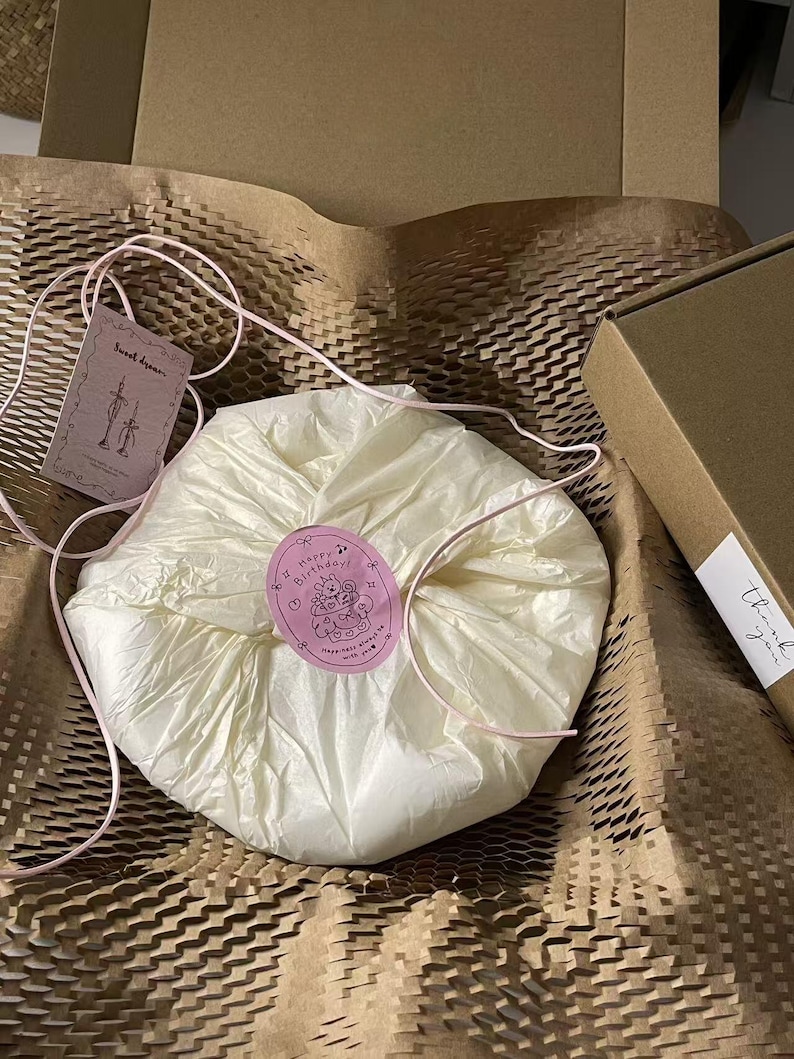 Peut inclure: Un paquet rond envelopp&eacute; dans du papier de soie cr&egrave;me, fix&eacute; par un autocollant rose repr&eacute;sentant un ours en dessin anim&eacute; avec le texte Happy Birthday. Une ficelle rose entoure le paquet, accompagn&eacute; d'une &eacute;tiquette marqu&eacute;e Sweet dream.
