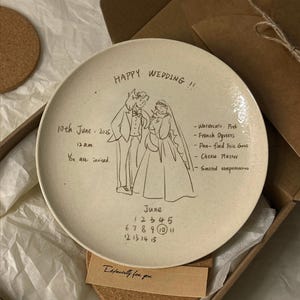 Peut inclure: Une assiette ronde en c&eacute;ramique blanc cass&eacute; avec un dessin au trait noir repr&eacute;sentant un couple de mari&eacute;s. Le texte inclut "HAPPY WEDDING !!", "10th June 2025", "12 a.m.", "You are invited.", un menu et un calendrier de juin avec le 10 entour&eacute;.
