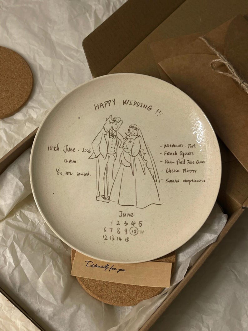 Peut inclure: Une assiette ronde en c&eacute;ramique blanc cass&eacute; avec un dessin au trait noir repr&eacute;sentant un couple de mari&eacute;s. Le texte inclut "HAPPY WEDDING !!", "10th June 2025", "12 a.m.", "You are invited.", un menu et un calendrier de juin avec le 10 entour&eacute;.