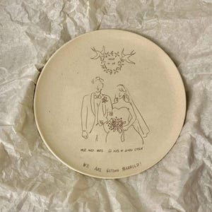 Peut inclure: Une assiette ronde en c&eacute;ramique cr&egrave;me mouchet&eacute;e avec une illustration au trait noir repr&eacute;sentant un couple en tenue de mariage. Au-dessus, une couronne florale porte la date 2025 12 25. En bas, le texte indique MR. AND MRS. LI XUE & ZHOU CHEN et WE ARE GETTING MARRIED!