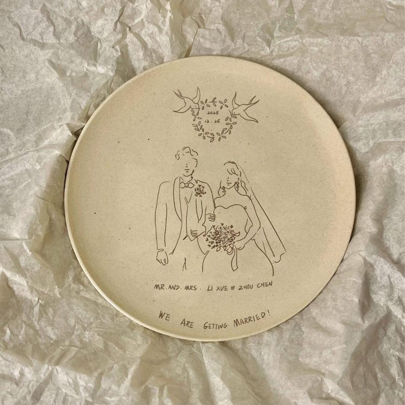 Peut inclure: Une assiette ronde en c&eacute;ramique cr&egrave;me mouchet&eacute;e avec une illustration au trait noir repr&eacute;sentant un couple en tenue de mariage. Au-dessus, une couronne florale porte la date 2025 12 25. En bas, le texte indique MR. AND MRS. LI XUE & ZHOU CHEN et WE ARE GETTING MARRIED!