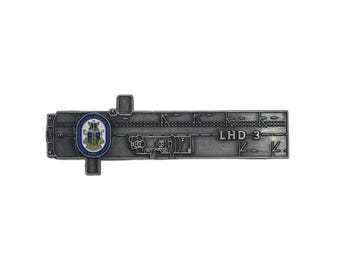 May include: A silver-toned metal lapel pin shaped like the flight deck of a naval ship. The pin features the text LHD 3, a small rectangular protrusion, and a circular emblem with blue, white, and green colors depicting a crest and the text USS KEARSARGE.