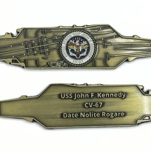 May include: A gold-colored metal challenge coin featuring a detailed outline of the USS John F. Kennedy aircraft carrier. The coin has the ship's name, "USS John F. Kennedy CV-67", and the motto "Date Nolite Rogare" on one side. The other side features a seal with the words "United States Ship John F. Kennedy" and "CV-67".
