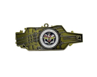 May include: A gold-colored metal pin in the shape of a naval aircraft carrier. The pin features the text "UNITED STATES SHIP JOHN F KENNEDY CV-67" around a central emblem. The number "67" is visible on the right side of the pin.