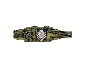 May include: A gold-colored metal pin in the shape of an aircraft carrier. The pin features detailed lines representing the ship's structure and a central shield with red and white stripes and a blue field with a white emblem.