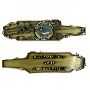 May include: A gold-colored metal challenge coin featuring a detailed outline of the USS Constellation aircraft carrier deck. The coin also features a circular emblem with the ship's name and the designation CV-64. The reverse side of the coin reads "USS Constellation CV-64 America's Flagship."