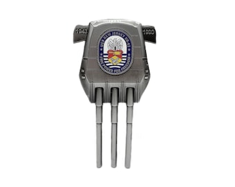 May include: A silver-colored metal comb-shaped object with the text "USS NEW JERSEY BB-62" and the years "1943" and "1990". The object has a crest with a shield and eagle design.