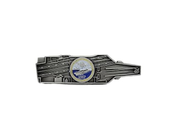 May include: A silver-colored metal pin shaped like an aircraft carrier. The pin features a circular emblem with a ship illustration and the words "Integrity, G.H.E.S." in blue and white. The pin has intricate details.