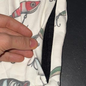 May include: White fabric with a black zipper and a pattern of colorful fishing lures. The lures are red, green, and blue.