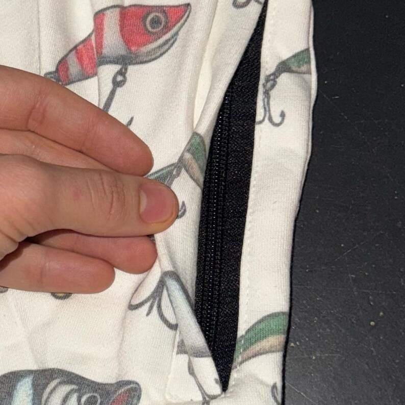 May include: White fabric with a black zipper and a pattern of colorful fishing lures. The lures are red, green, and blue.